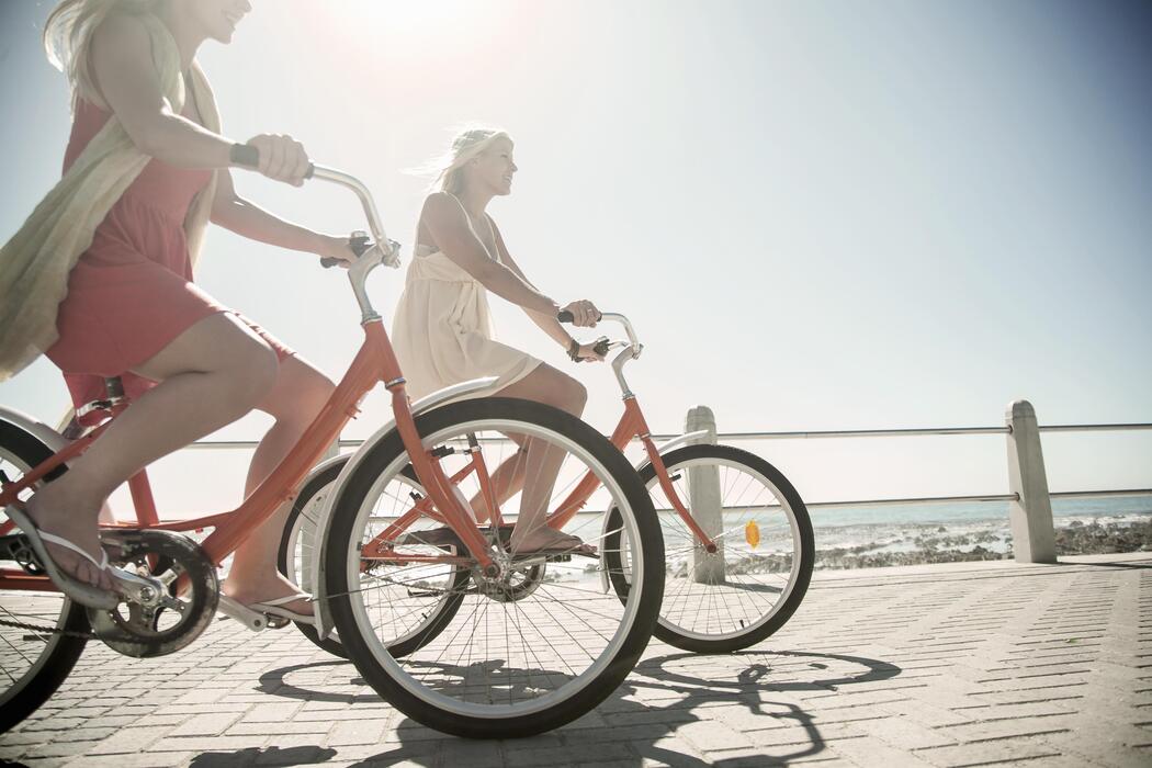 Two women in summer dresses riding orange cruiser bicycles along a sunny coastal boardwalk beside the ocean